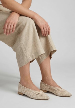 Kobieta nosząca beżowe plecione baleriny z zaostrzonym noskiem i luźne, jasne lniane spodnie, siedząca z rękami spoczywającymi na kolanach na jednolitym tle.