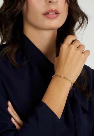 Bracciale in oro con elementi circolari e a catena alternati e testurizzati. Indossato al polso, abbinato a un indumento blu scuro.