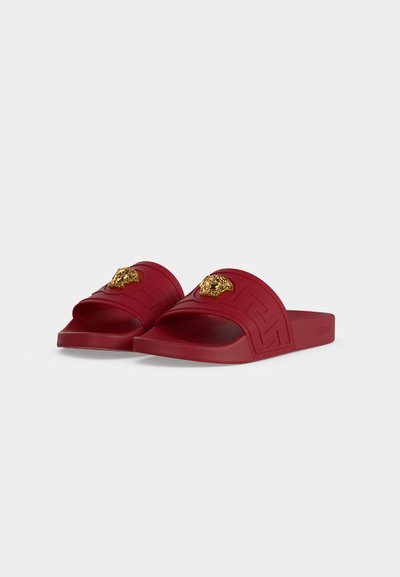 Sandales rouges à enfiler avec ornements en tête de lion dorés et motifs géométriques embossés sur les brides, sur fond blanc uni.