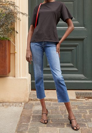 Osoba ubrana w czarną bluzkę z krótkim rękawem, krótkie niebieskie dżinsy, czarne sandały na obcasie z paskami, niosąca czerwoną torebkę na ramię, stojąca na kamiennej nawierzchni.