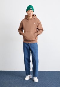 Ein junger Mann trägt einen braunen Hoodie, blaue Jeans, weiße Sneakers und eine grüne Mütze. Er steht mit den Händen in der Hoodie-Tasche vor einer schlichten Wand.