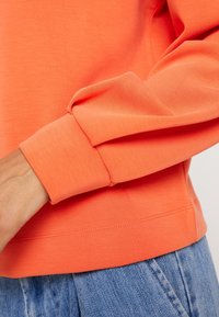 Oranger Pullover mit glatter Textur, ausgestattet mit gerippten Bündchen und Saum. Das Design umfasst einen lockeren Schnitt und sichtbare Nähte.