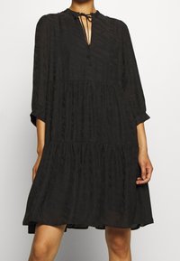 Vestido negro de capas, confeccionado con una tela ligera y texturizada. Presenta un escote con forma de llave y lazos, además de mangas tres cuartos. Silueta fluida.