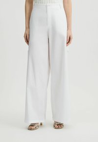 Weiße, weit geschnittene Hosen mit glatter Textur, hoher Taille und vorderem Knopfverschluss; kombiniert mit metallischen Riemchensandalen.