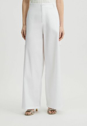Pantaloni bianchi a gamba larga con una texture liscia, vita alta e chiusura frontale con bottone; abbinati a sandali metallici con strappi.