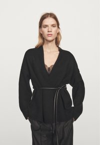 Černý pletený cardigan s výstřihem do V, širokými rukávy a látkovým páskem v pase. Má dvě přední kapsy a strukturovaný povrch.