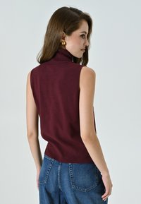 Burgunderfarbener ärmelloser Rollkragenpullover mit geripptem Saum; weiche Stricktextur; kombiniert mit hochtaillierten Jeans, die eine lockere Passform zeigen.