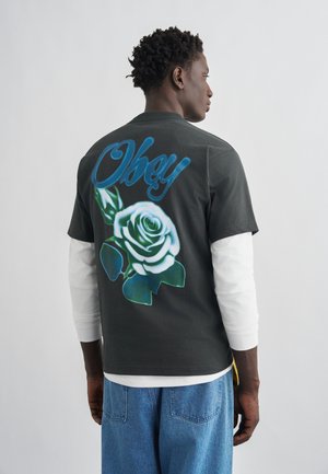 Άνδρας που φοράει πολυεπίπεδη λευκή μακρυμάνικη μπλούζα και μαύρο T-shirt με μπλε κείμενο "Obey" και μπλε γραφικό λουλούδι τριαντάφυλλο στο πίσω μέρος, στέκεται μπροστά από απλό φόντο.