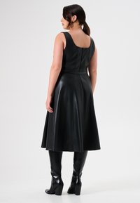 Zwarte leren jurk met een getailleerde bovenkant en een uitlopende rok, voorzien van een ronde rug en een gladde textuur. Gepaard met knie-hoge leren laarzen.