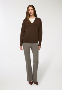 Brązowy, oversize, sweter z dekoltem w serek wykonany z miękkiego materiału, w połączeniu z jasnoszarymi, dopasowanymi spodniami. Całość uzupełniają brązowe szpilki o wydłużonym czubku.