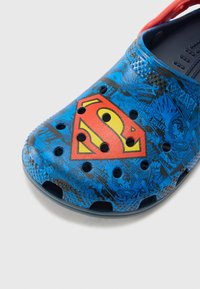 Modré Crocs se Supermanovým logem; texturovaný povrch, otevřené díry pro ventilaci a červený popruh. Obsahují grafické detaily ve stylu komiksu.
