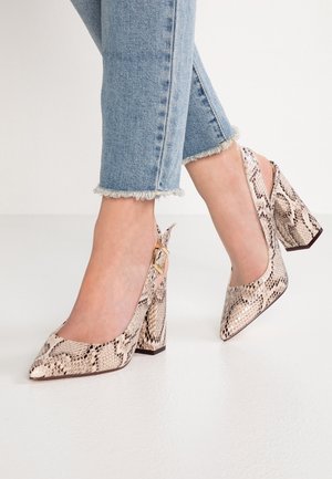 Des pieds portant des chaussures slingback à motif serpent, à bouts pointus et talons blocs, associés à un jean bleu clair à ourlet effiloché sur un sol blanc.