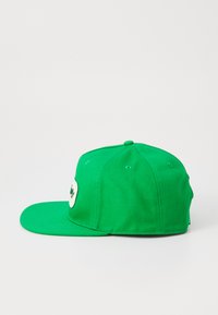 Green snapback cap featuring a rounded crown, stiff bill, and an embroidered logo patch on the front. Made from textured cotton material.