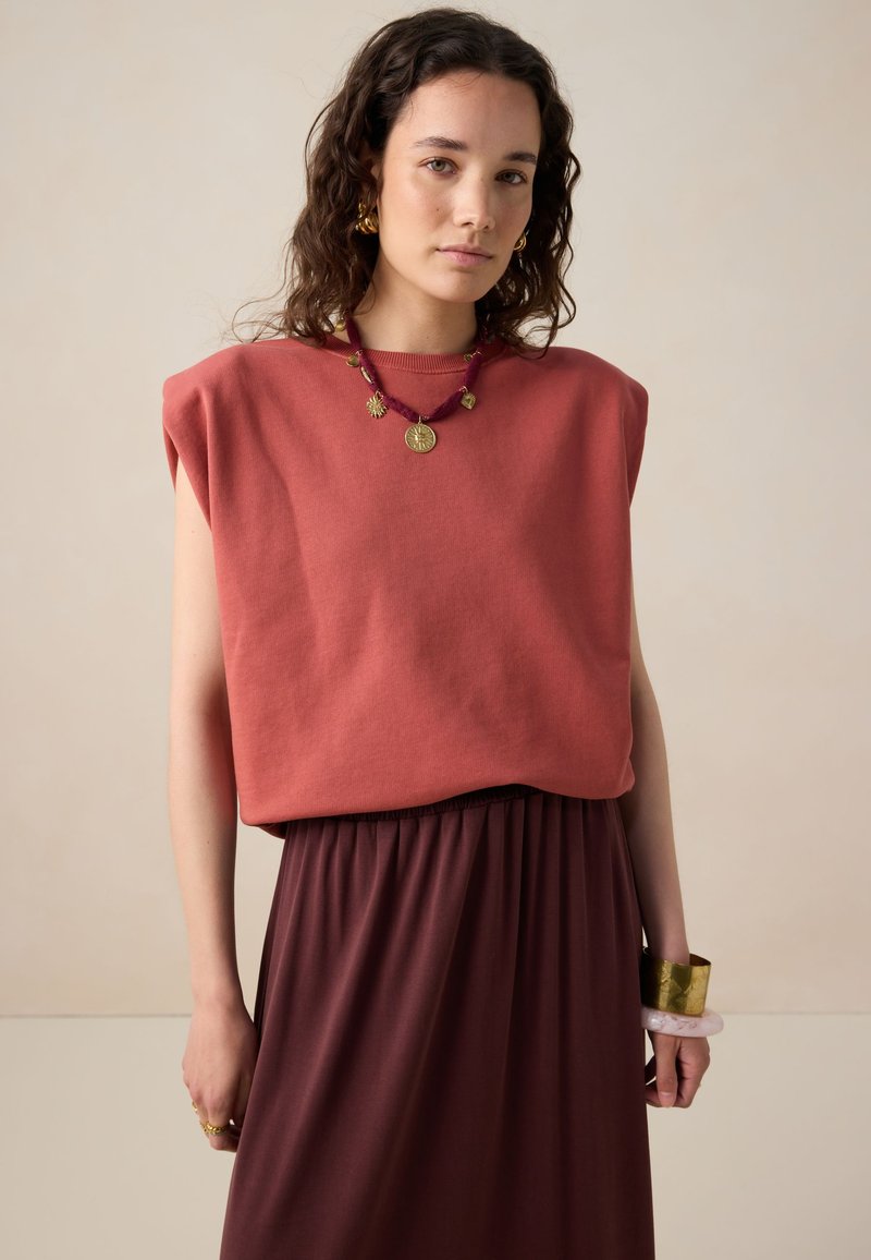 Haut sans manches corail en tissu doux, associé à une jupe rouge foncé. Accessoirisé avec un collier et un bracelet en perles dorées et colorées.