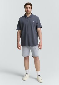Dunkelgraues Poloshirt mit kurzen Ärmeln, ausgestattet mit einem Kragen und einem Logo. Kombiniert mit hellgrauen Shorts und weißen Sneakers mit beigen Akzenten.