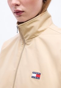 Άτομο που φοράει μπεζ ζακέτα με φερμουάρ και ψηλό γιακά, με την τσέπη λογότυπου Tommy Jeans στο στήθος, φαίνονται τα χείλη και το κάτω μέρος του προσώπου.