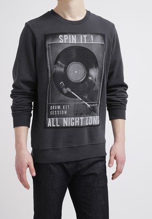 Osoba nosząca czarną bluzę z grafiką winylowej płyty i gramofonu oraz tekstem "Spin it! Drum kit session all night long."