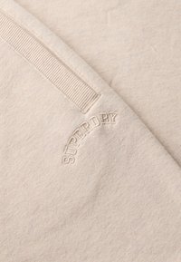 Tessuto di felpa beige con una texture morbida, caratterizzato da un accento a coste e logo "SUPERDRY" in rilievo in formato curvo su un lato.