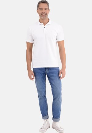 Homme souriant portant un polo blanc, un jean bleu retroussé et des baskets blanches, debout devant un fond clair uni.