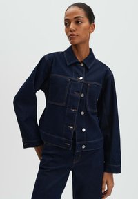 Giacca di denim blu scuro con colletto, quattro tasche anteriori e bottoni in metallo argentato. Presenta cuciture arancioni a contrasto e una vestibilità comoda.