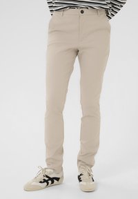 Pantalons en mélange de coton beige à coupe slim, dotés d'une fermeture à bouton et de poches avant, associés à des baskets beige et noires.