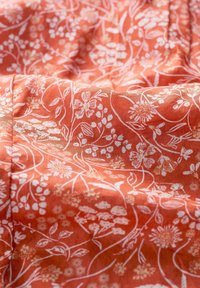 Terrakotta-tyg med vitt blommönster; har intrikata blommor och vinrankor med en texturerad, något rynkad yta.
