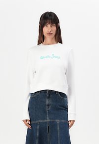 Baltas džemperis su turkio spalvos "Guess Jeans" logotipu priekyje, su apvaliu iškirpimu ir raukšlėtais rankogaliais, derinamas su tamsiomis džinsinėmis sijonais.
