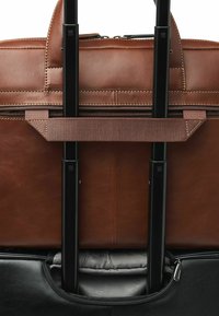 Braune Ledertasche mit strukturierter Oberfläche, schwarzer Beschläge und einem Reißverschlussfach, entwickelt, um sicher an Rollgepäck befestigt zu werden.