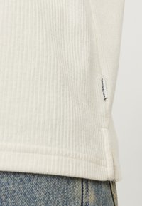 Camiseta de color crema de rayas con superficie texturizada, presenta una abertura lateral y una pequeña etiqueta en el dobladillo. Combinada con vaqueros de mezclilla azules.