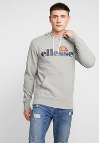 Grijze hoodie van een katoenmix met een voorkantzak en witte koorden. Bevat een groot "ellesse" logo in donkerblauw met een oranje accent.