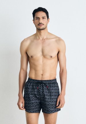Shorts de bain noirs avec un motif logo répété, une taille à cordon avec des accents rouges et une coupe décontractée. Texture de tissu lisse.