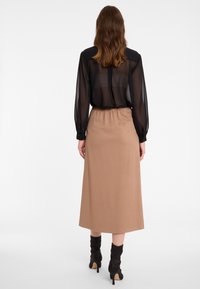 Schwarze transparente Bluse mit langen Ärmeln und geknöpften Manschetten, kombiniert mit einem mittellangen beigen Rock mit elastischem Bund. Sichtbare schwarze Stiefel mit Absatz.