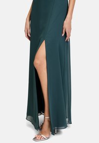 Grünes, schwingendes Maxikleid mit einem Oberschenkel-Schlitz. Der Stoff ist glatt, während silberne, verstellbare Absätze das Outfit ergänzen.