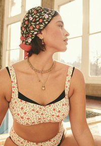 Floral bikini top with red and pink patterns on a light background, paired with a black bottom, gold chain necklace, and a patterned scarf.