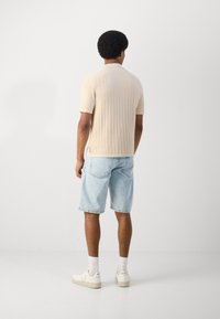Beige ribbed knit short-sleeve polo shirt paired with light blue denim shorts. Footwear is white sneakers. Minimalist style, plain background.