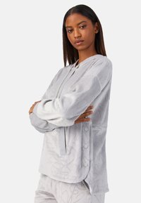 Sudadera gris con capucha y patrón texturizado sutil, cierre frontal con cremallera, puños acanalados y botones laterales. Tela suave con ajuste relajado.