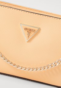 Portefeuille en texture pêche présentant une plaque en métal triangulaire gravée "GUESS", orné d'une chaîne argentée le long du bord.