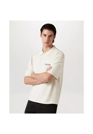Jeune homme aux cheveux foncés portant un polo blanc à manches courtes avec le logo « BELSTAFF », les bras croisés, debout devant un fond blanc uni.