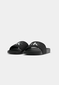 Sandales slides noires avec larges brides en tissu ornées de logos blancs Calvin Klein "CK", vue en angle sur un fond clair uni.