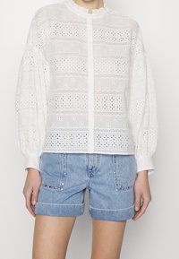 Blusa bianca con intricati motivi a taglio, scollatura rotonda e maniche a sbuffo, abbinata a pantaloni in denim con dettagli di borchie.