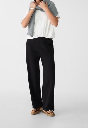 Vrouw draagt zwarte wijde broek, wit T-shirt met korte mouwen, grijze trui over de schouders gedrapeerd en sneakers met luipaardprint.