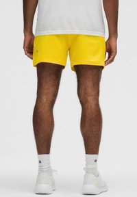Gula idrottsshorts med en slät textur, över knä längd, kombinerat med vita sneakers och strumpor, sedd bakifrån.