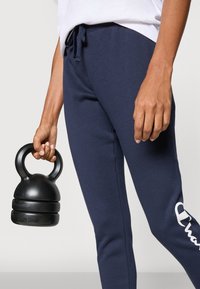 Mörkblå sweatpants med elastisk midja och dragband, med en vit logotyp. En svart kettlebell hålls i en hand.