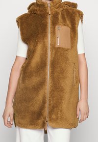 Brązowa kamizelka z polaru z kapturem, z przednim zamkiem błyskawicznym i kieszenią na piersi wykonaną z kontrastowego, cielistego materiału. Gładka faktura z puszystym, gęstym włosiem.