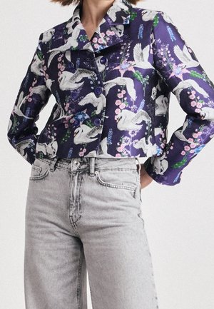 Femme portant une veste courte violette avec un motif de cygnes blancs et de fleurs roses, associée à un jean taille haute gris clair.