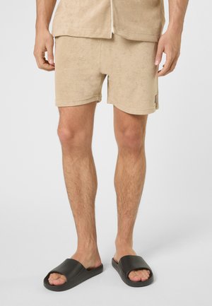 Uomo che indossa pantaloncini in spugna beige e camicia coordinata con sandali neri a fascia, in piedi davanti a uno sfondo bianco.