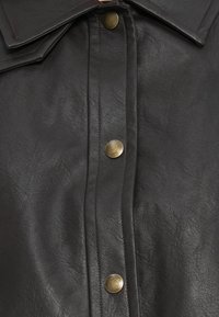 Chemise en cuir noir avec une finition texturée, dotée d'une patte de boutonnage à l'avant et de boutons-pression en laiton, accentuée par un large col.