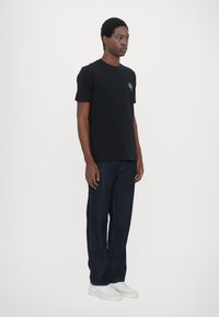 Camiseta de algodón negra con mangas cortas, con un gráfico en el lado izquierdo del pecho. Combinada con jeans de mezclilla azul oscuro y zapatillas blancas.