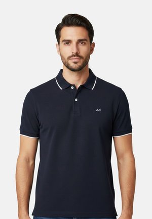 Uomo che indossa una polo blu navy con bordo bianco su colletto e maniche, in piedi contro uno sfondo grigio chiaro semplice.