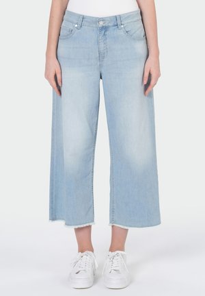 Hellblaue, weit geschnittene Cropped-Jeans mit rohen Säumen, getragen mit weißen Sneakern und einem weißen Oberteil, von der Taille bis zu den Füßen gezeigt.